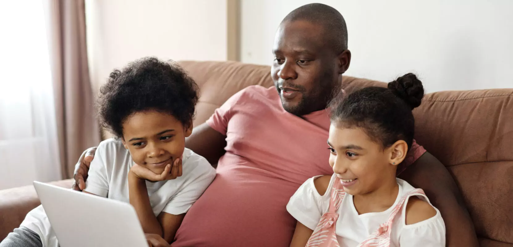 a dad and his two kids sit on a couch looking at the computer