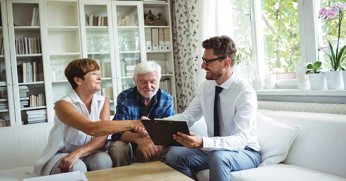 a couple and an advisor sit on a couch and shake hands while looking at a clip board