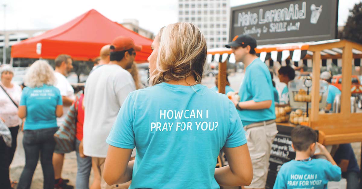 a woman wearing a shirt that says "how can I pray for you?" in a crowd of people