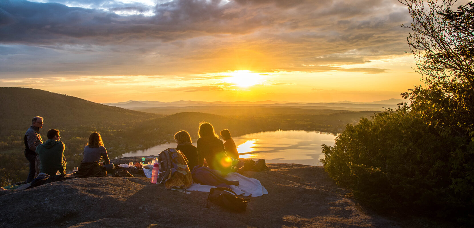 a group of friends sitting on top of a mountain overlooking a lake at sunset